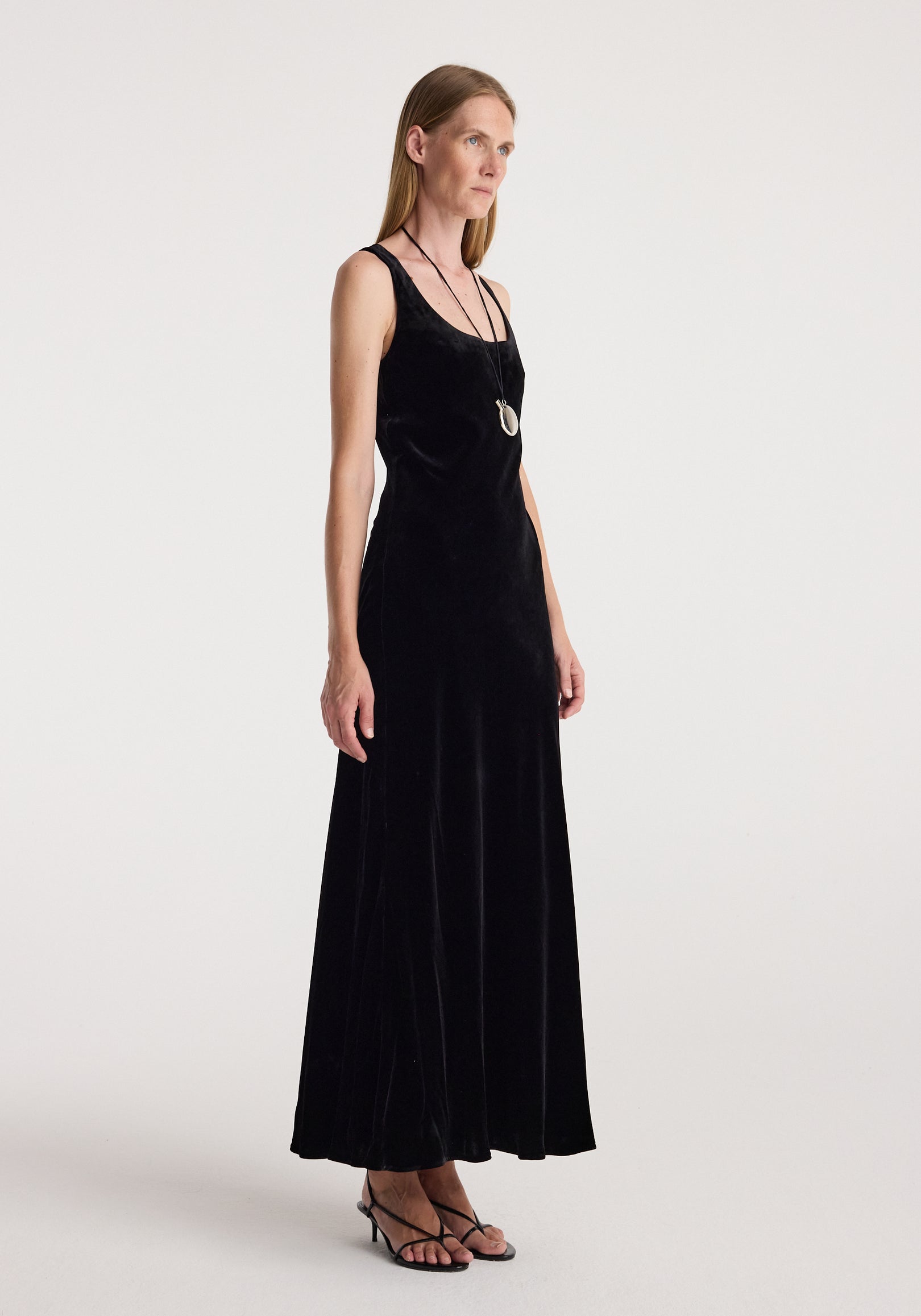 Woman wearing a long black scoop-neck velvet dress | black with thin straps, paired with strappy black heels and a pendant necklace standing against a plain background.