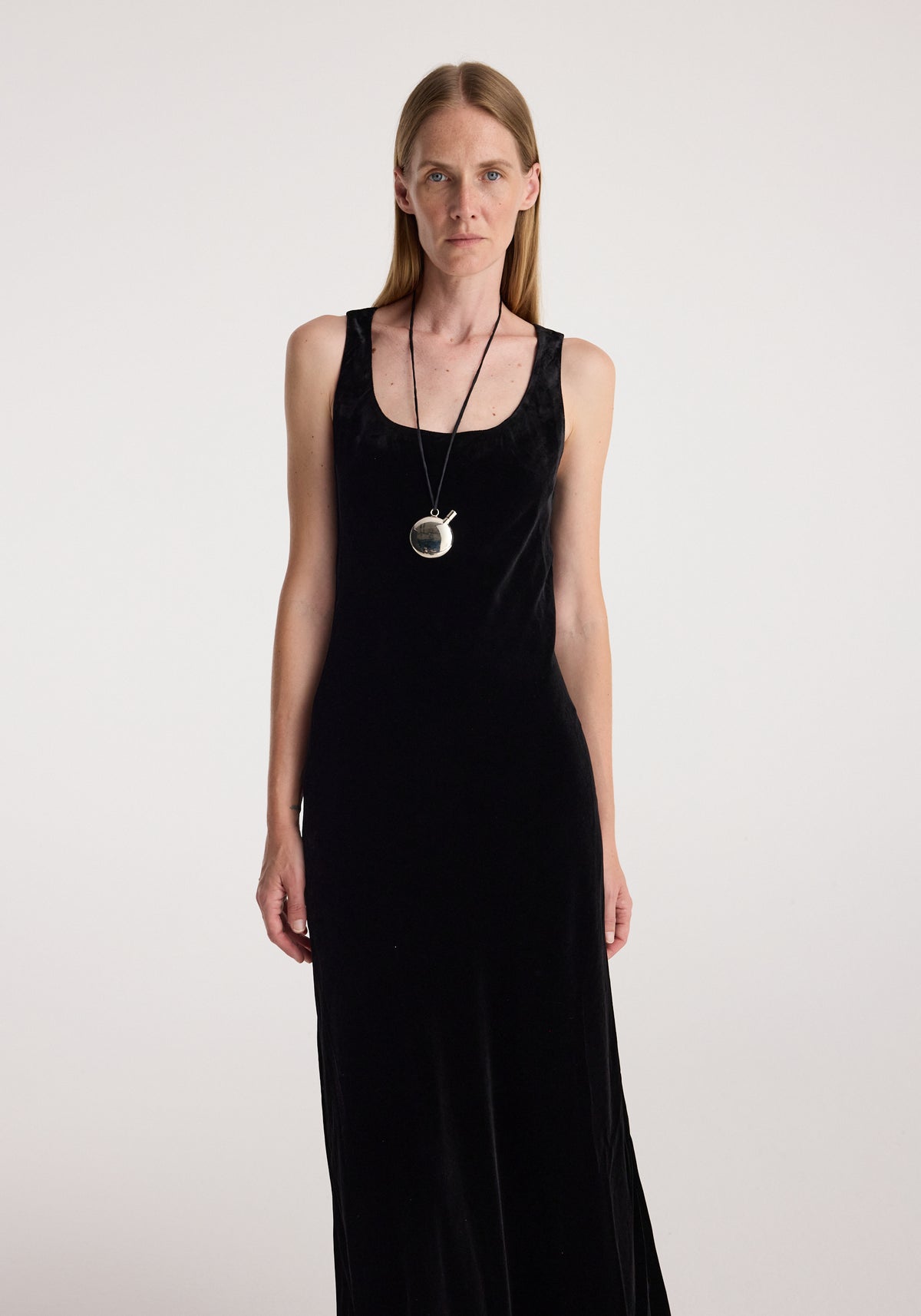 Woman wearing a sleeveless black scoop-neck velvet dress standing against a plain light background, accessorized with a long pendant necklace, showcasing the Scoop-neck velvet dress | black.
