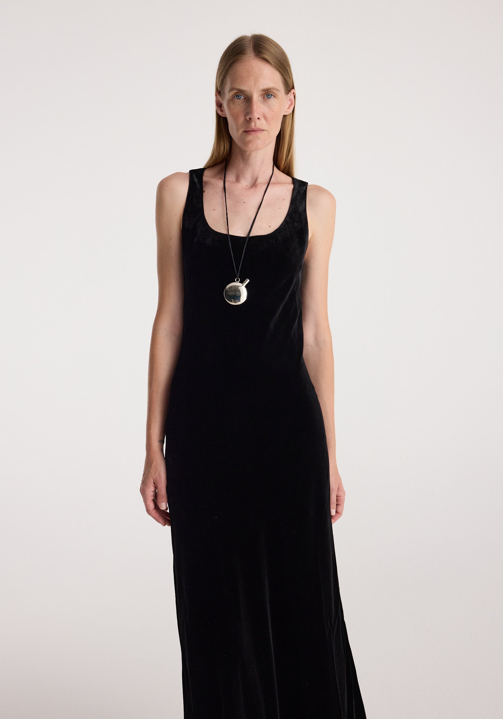 Woman wearing a black sleeveless dress and a long silver chain with a Silver perfume bottle necklace | silver pendant around her neck against a plain background.