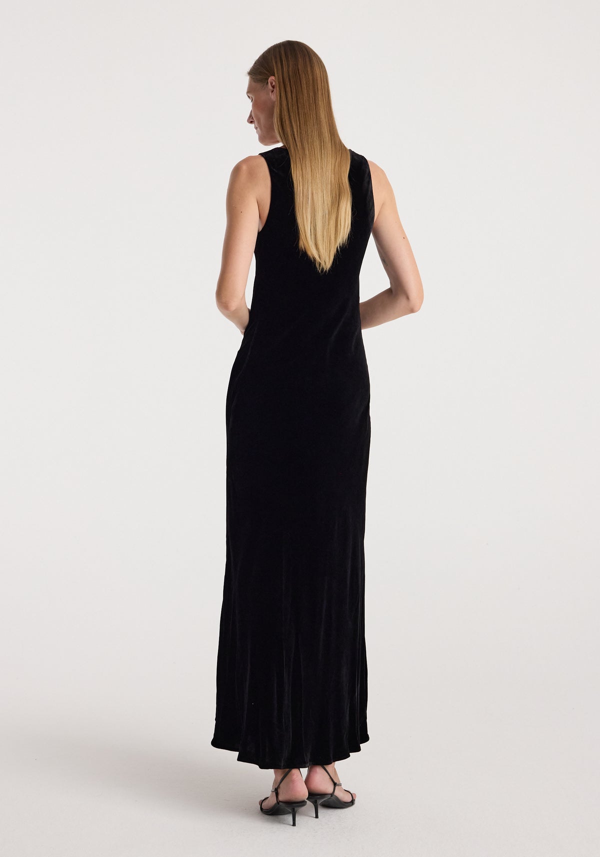 Woman wearing a long black scoop-neck velvet dress | black, shown from the back with sleeveless design and mid-heel sandals.