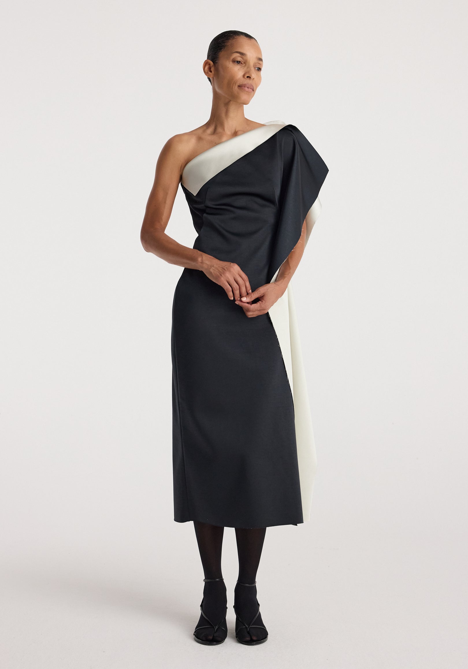 Woman wearing a sculptural double-satin off-shoulder dress | black/white, standing with one arm folded and looking to the side against a plain light background.