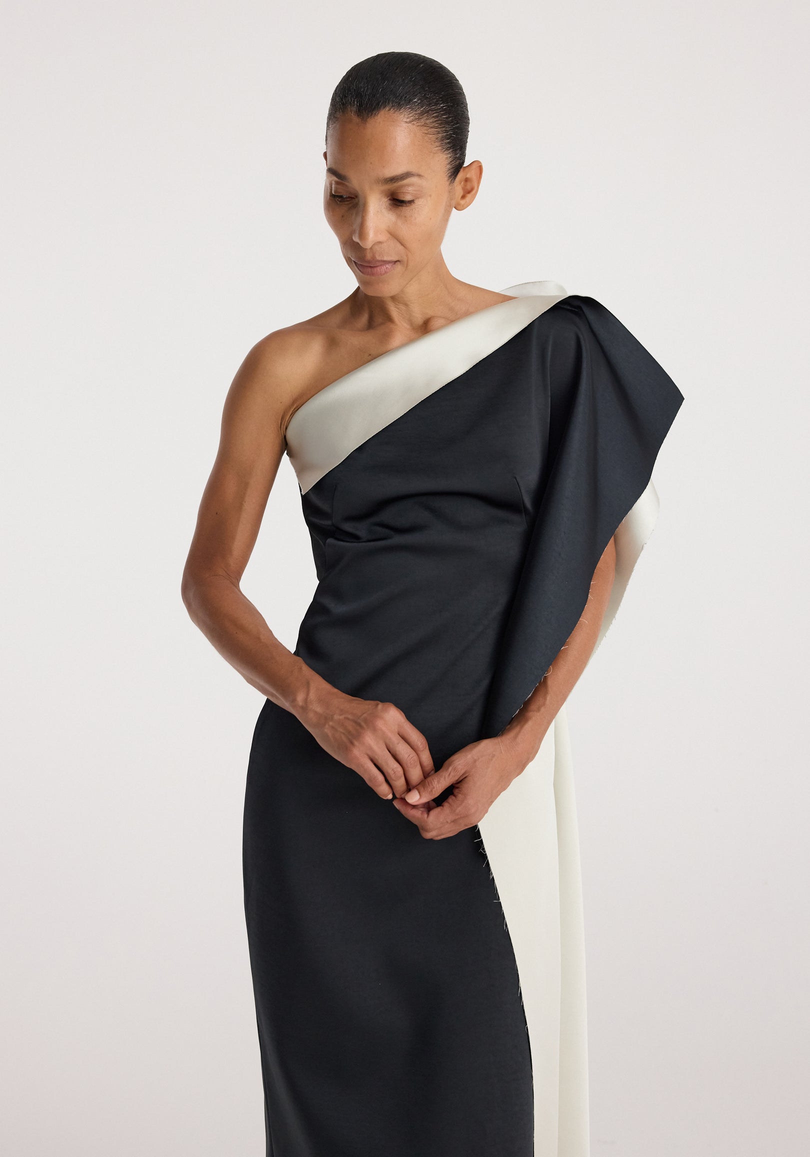 Woman wearing a sculptural double-satin off-shoulder dress | black/white with an asymmetrical design and draped sleeve detail against a plain background.
