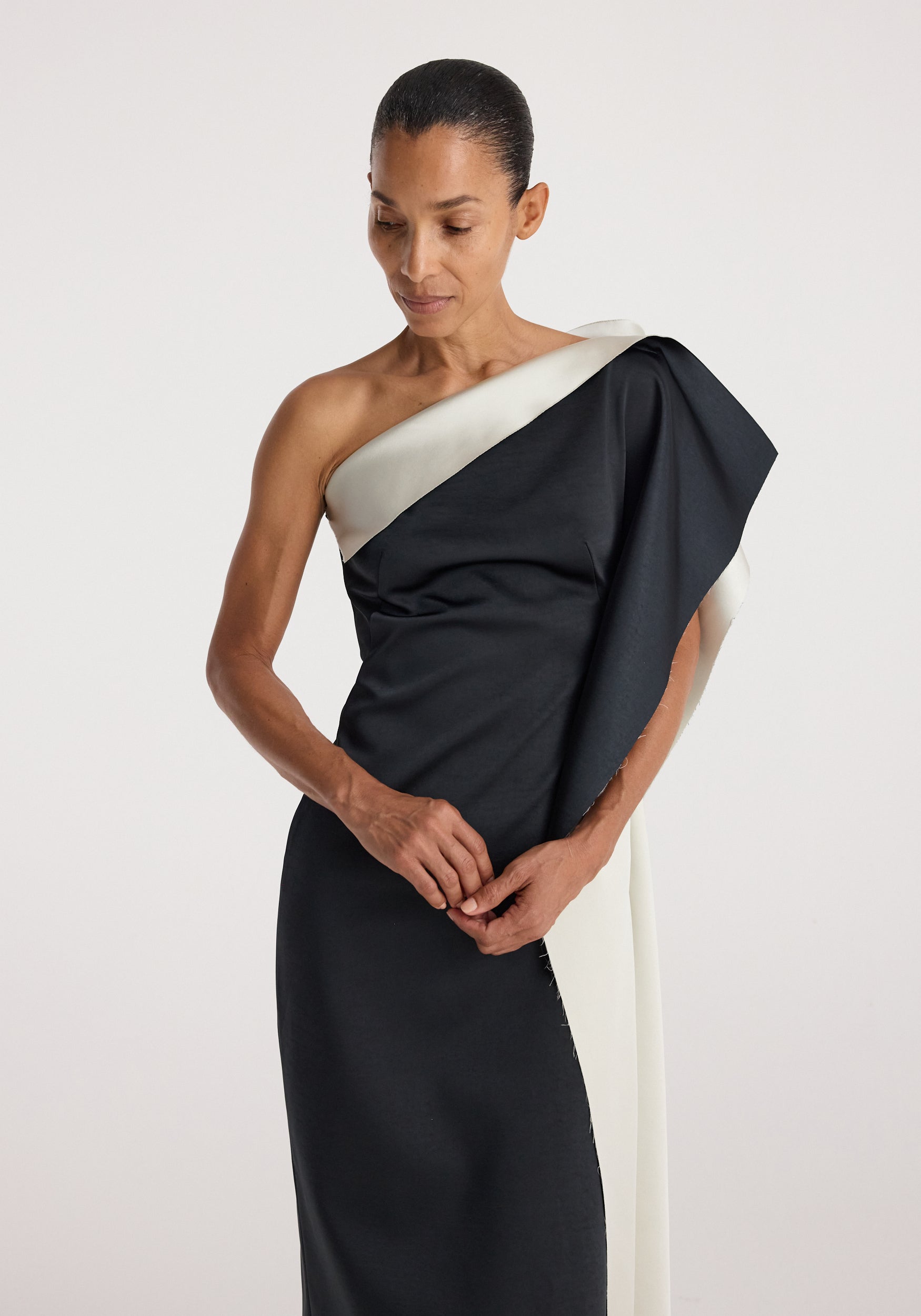 Woman wearing a sculptural double-satin off-shoulder dress | black/white with an asymmetrical design and draped sleeve detail against a plain background.