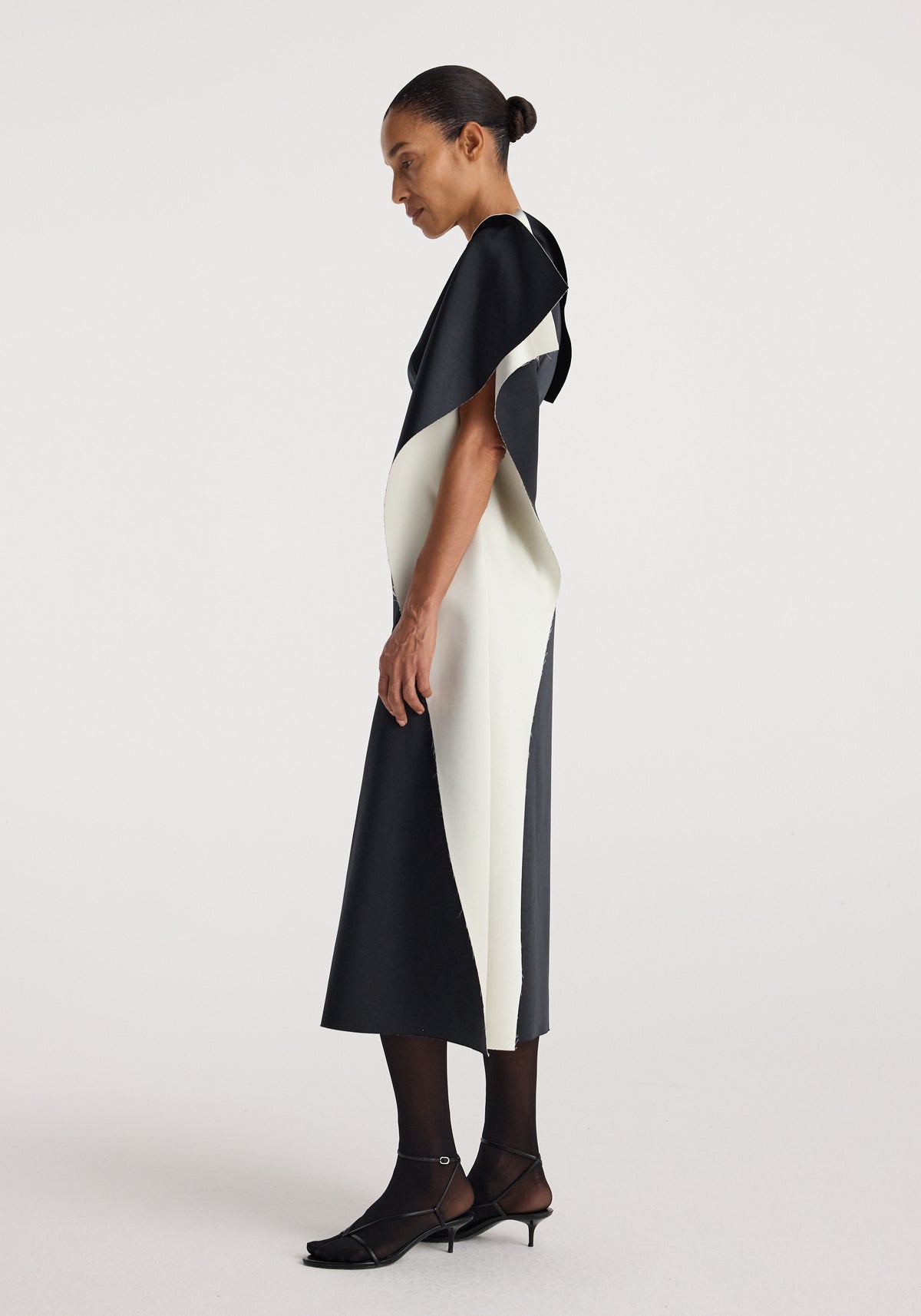 Profile view of a model wearing a sculptural double-satin off-shoulder dress | black/white featuring a sleek, color-block design with black and white panels and elegant draping.