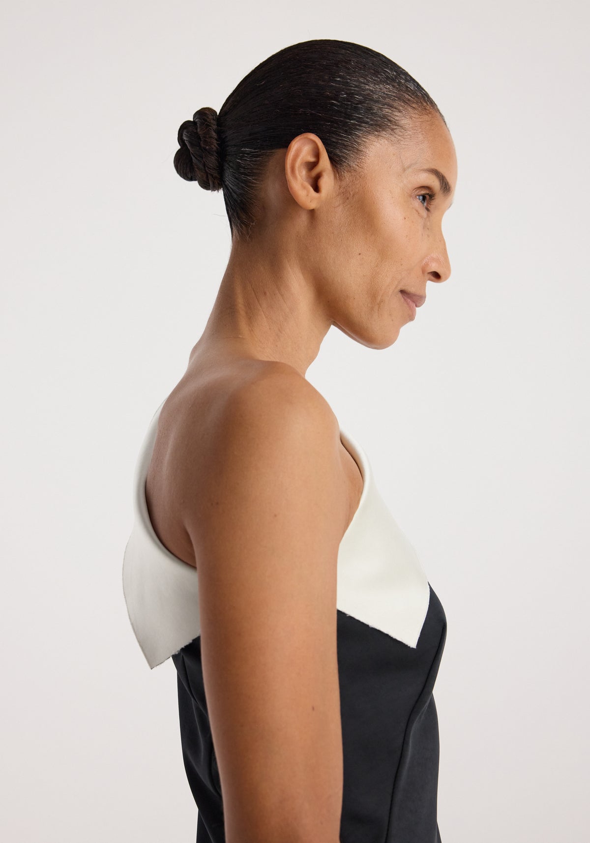 Side profile of a woman wearing a sculptural double-satin off-shoulder dress | black/white with a sleek hairstyle against a plain light background.