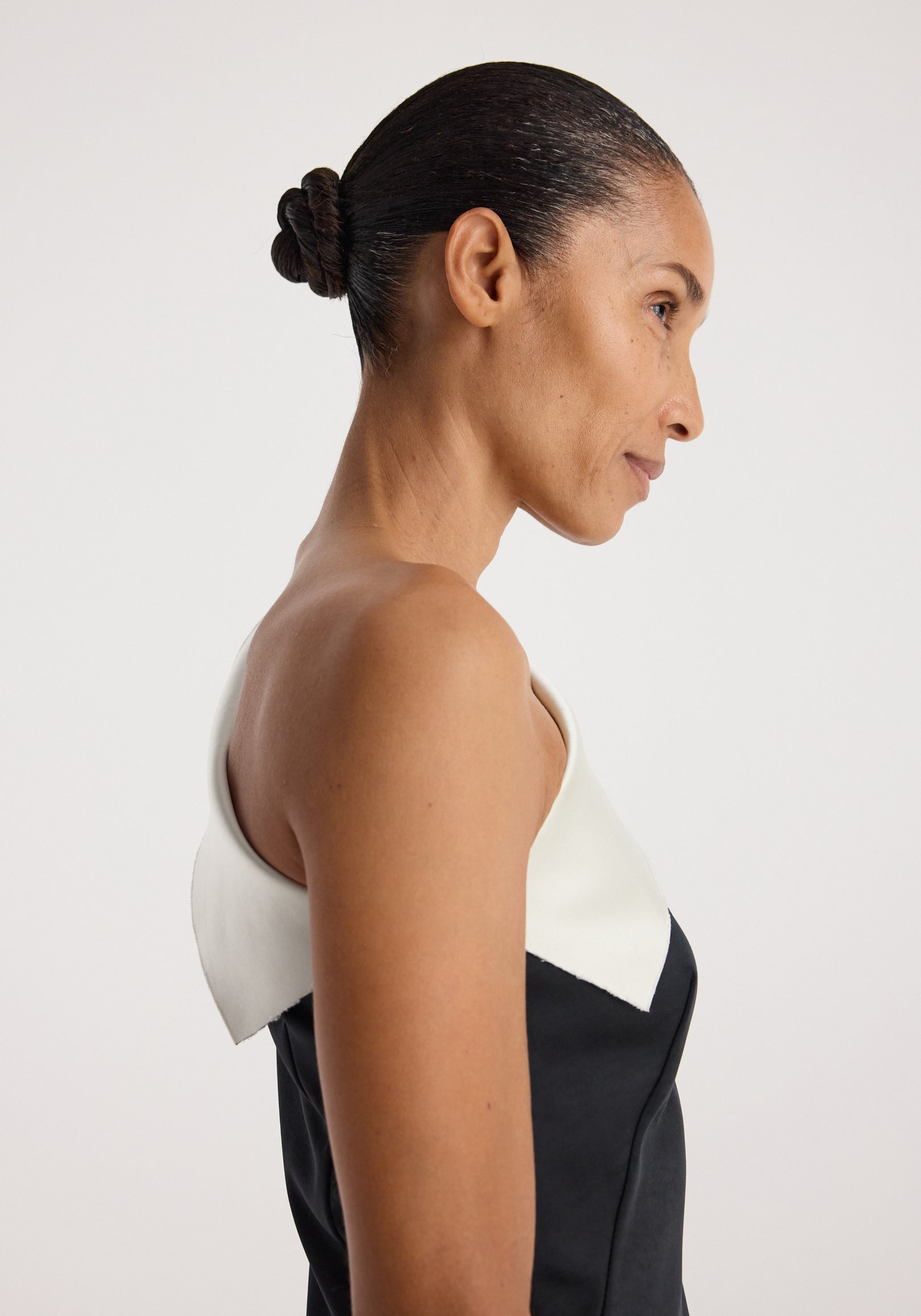 Side profile of a woman wearing a sculptural double-satin off-shoulder dress | black/white with a sleek hairstyle against a plain light background.