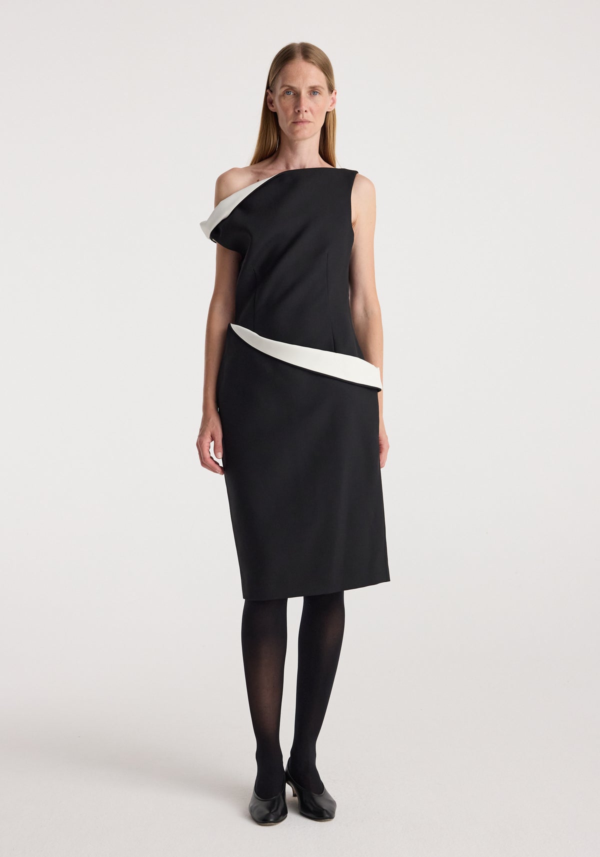 Woman wearing a Sculptural off shoulder dress | black with white sculptural details around the neckline and waist, paired with black tights and black shoes on a plain background.