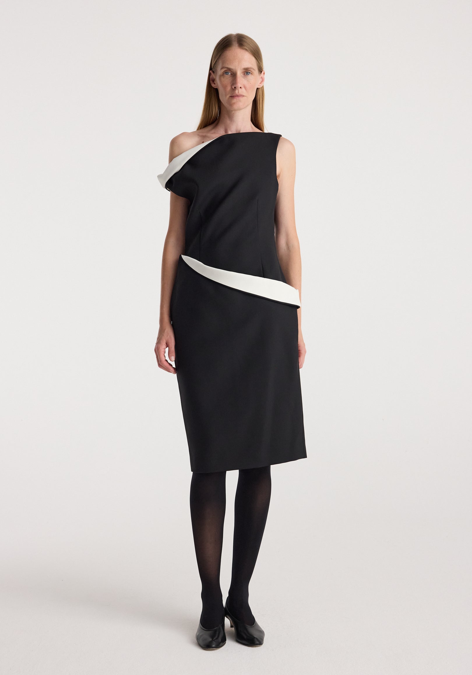 Woman wearing a Sculptural off shoulder dress | black with white sculptural details around the neckline and waist, paired with black tights and black shoes on a plain background.