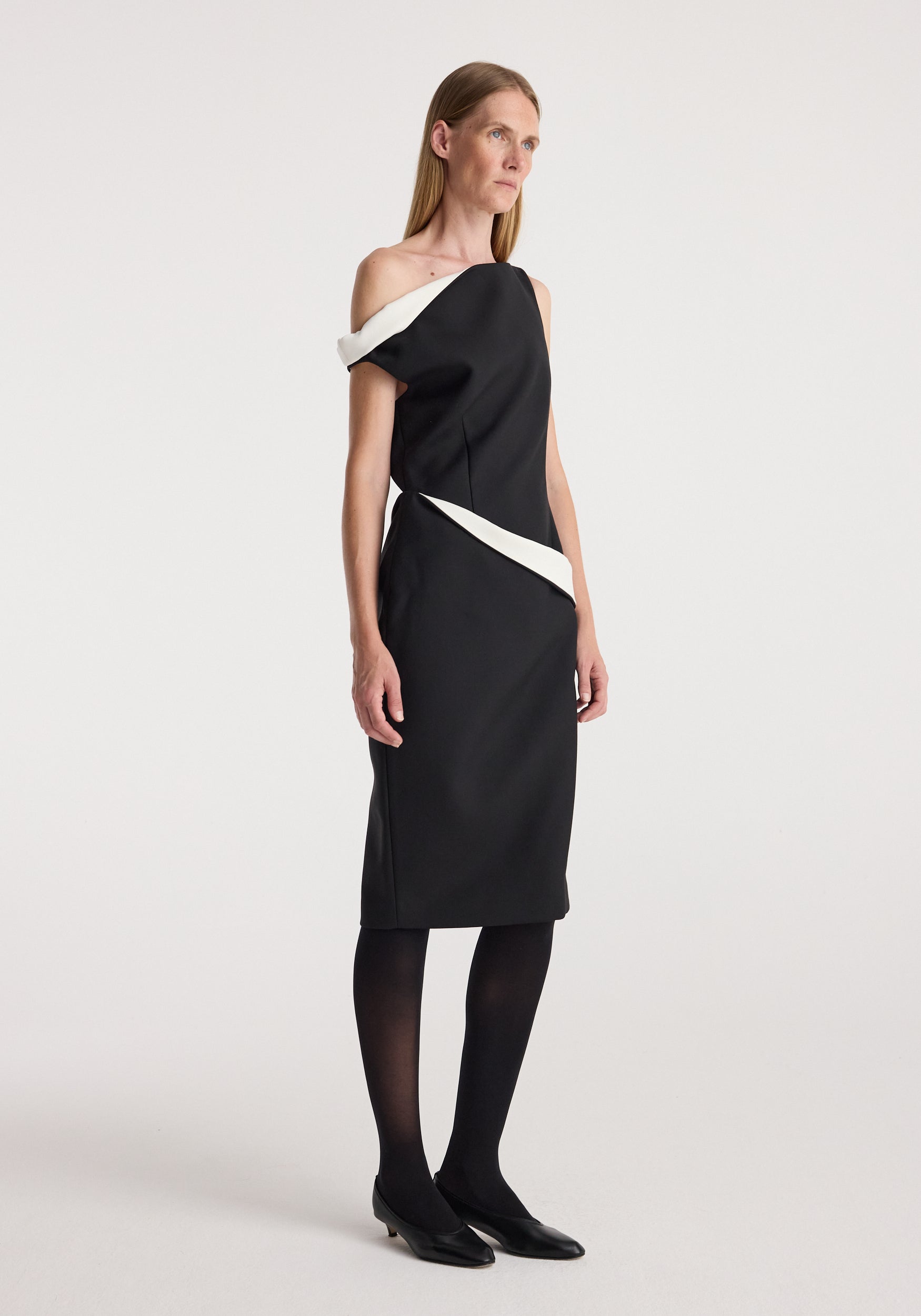 Female model wearing a Sculptural off shoulder dress | black with white sculptural details, paired with black tights and black low-heeled shoes against a plain background.