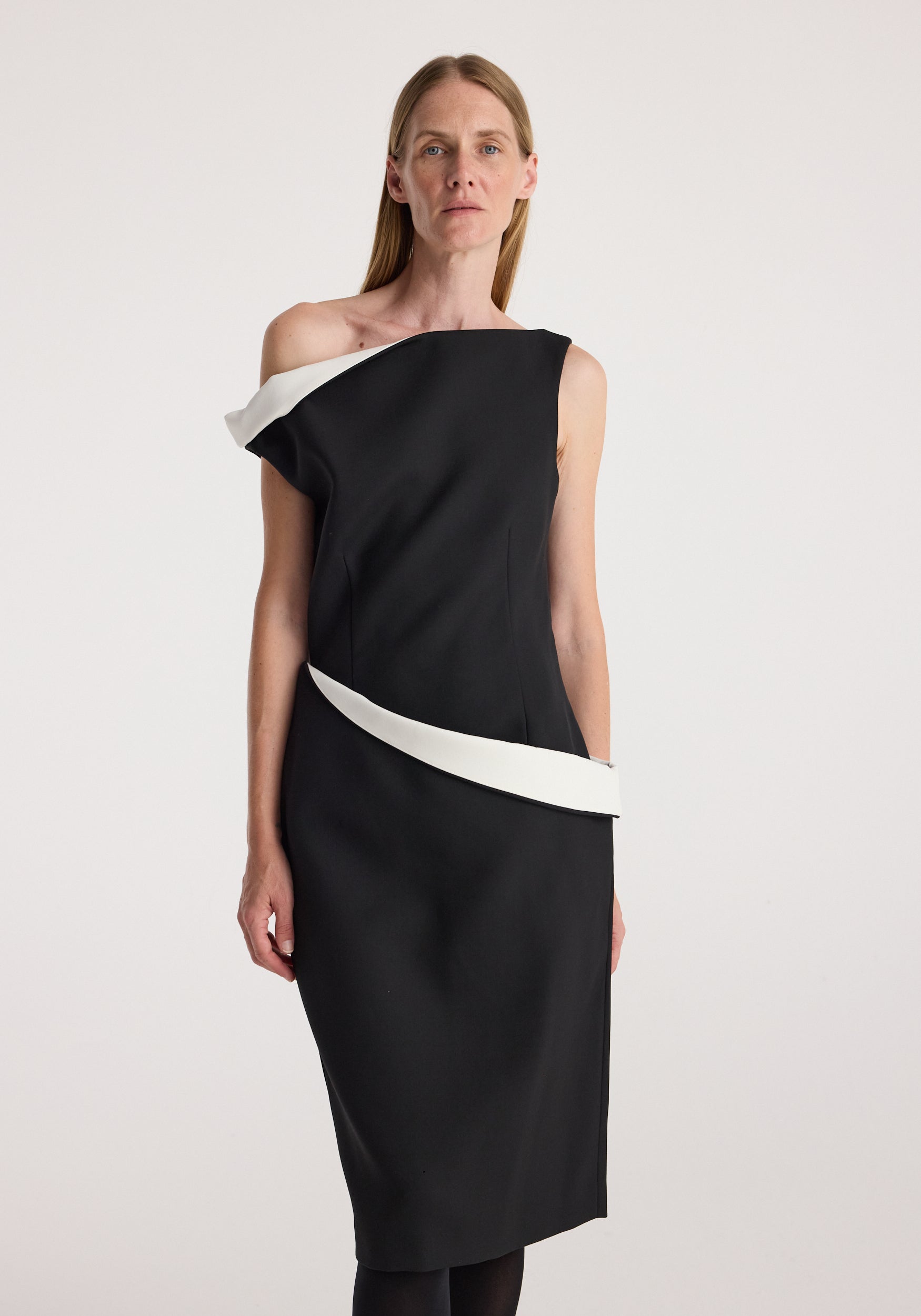 Woman wearing a black Sculptural off shoulder dress | black with asymmetrical white accents along the shoulder and waist, standing against a plain light background.