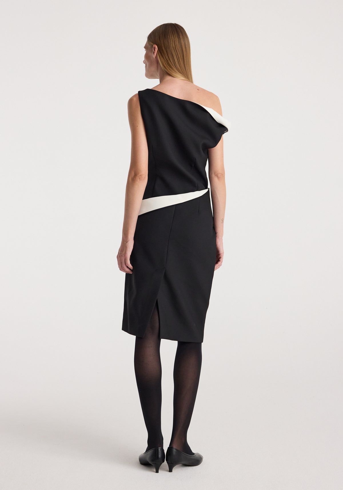 Woman wearing a Sculptural off shoulder dress | black with a white accent detail, shown from the back, styled with black tights and black heels against a plain background.