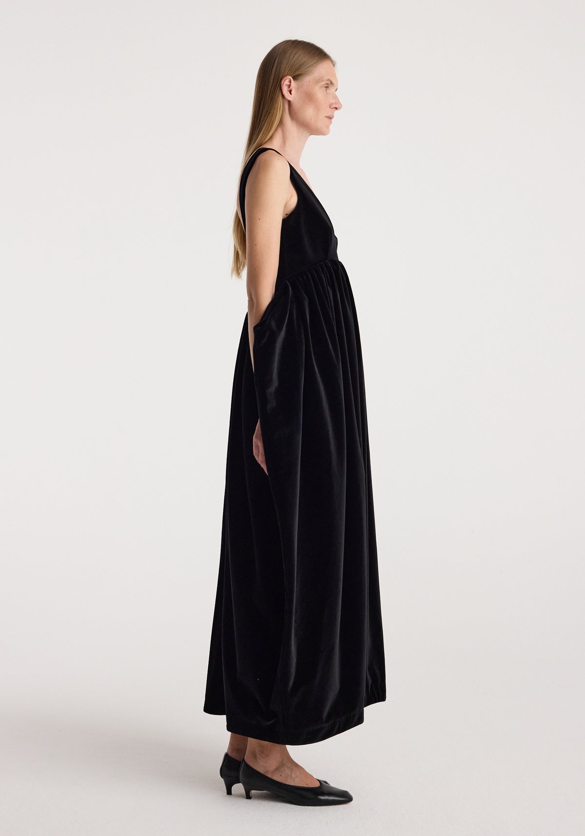 Side view of a model wearing the Sculptural volume velvet dress | black, a long flowing black velvet dress with a gathered waist and sleeveless silhouette.