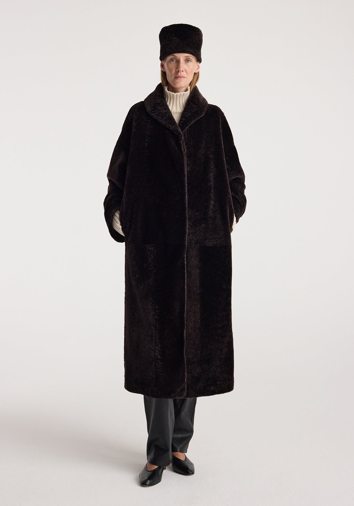 Shearling hat | dark chocolate worn by a model with a long dark shearling coat, cream knit sweater, dark trousers, and black shoes against a light background
