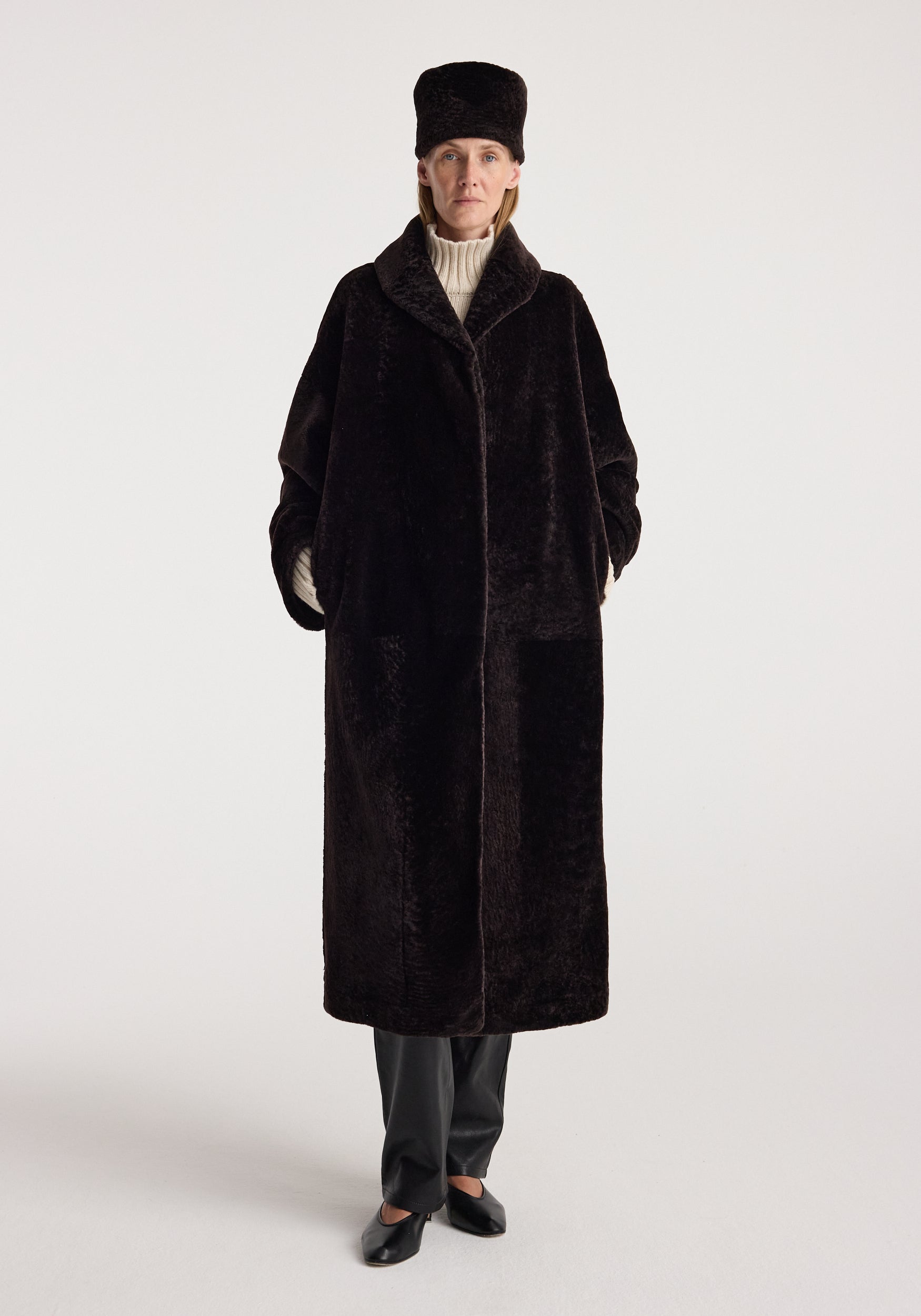 Shearling hat | dark chocolate worn by a model with a long dark shearling coat, cream knit sweater, dark trousers, and black shoes against a light background