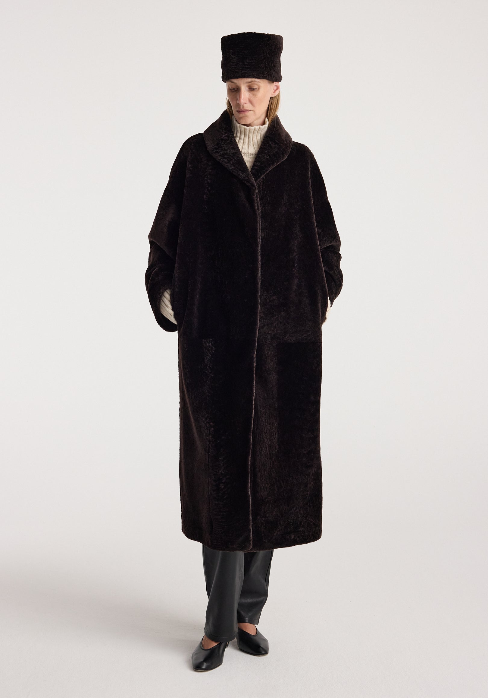Shearling hat | dark chocolate worn by a model in a dark brown shearling coat, styled with a cream turtleneck, trousers, and black shoes against a light background