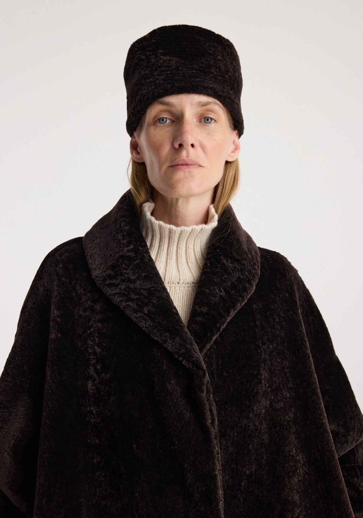 Shearling hat | dark chocolate worn by a model with a dark brown coat and cream ribbed turtleneck against a light background