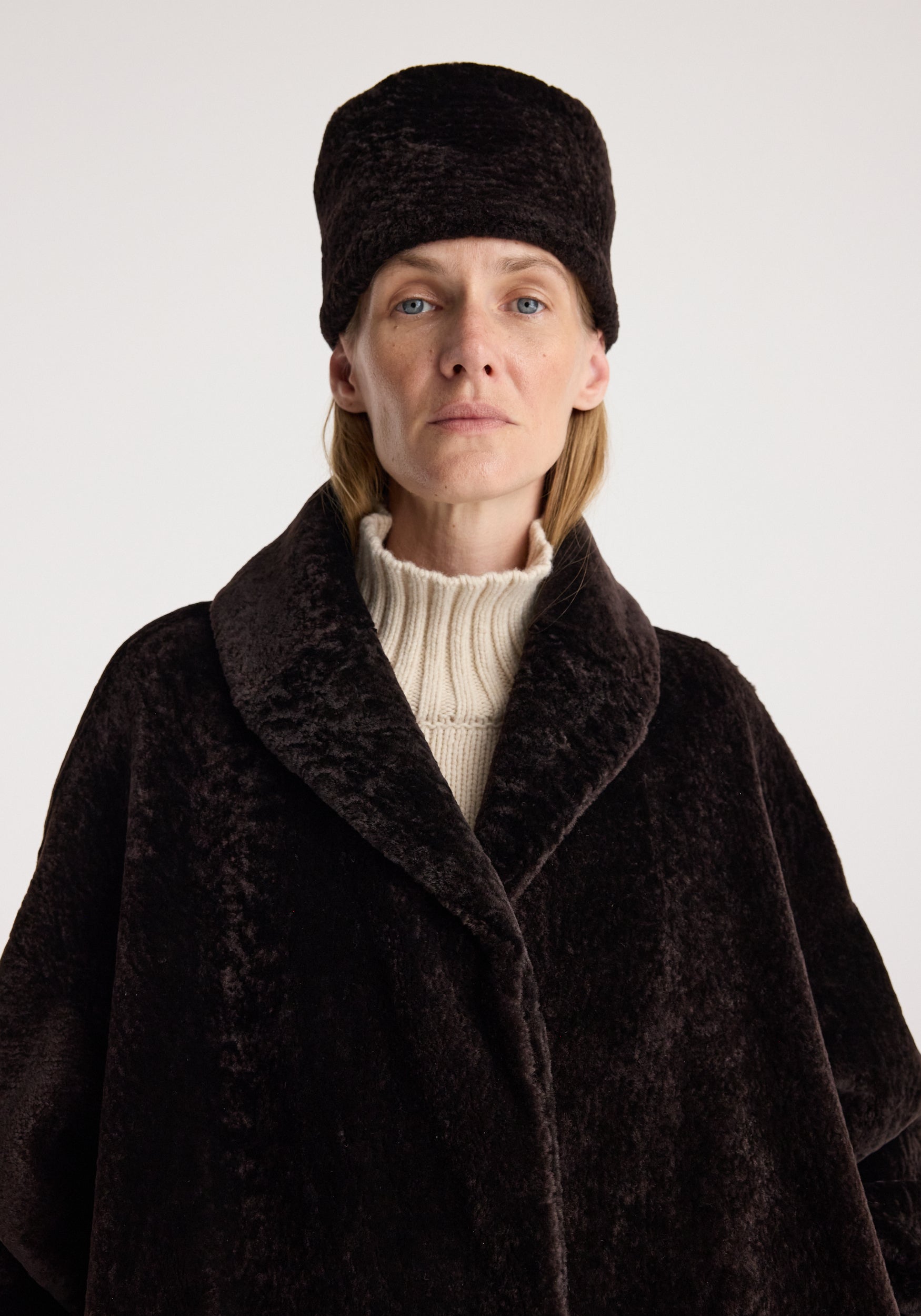 Shearling hat | dark chocolate worn by a model with a dark brown coat and cream ribbed turtleneck against a light background