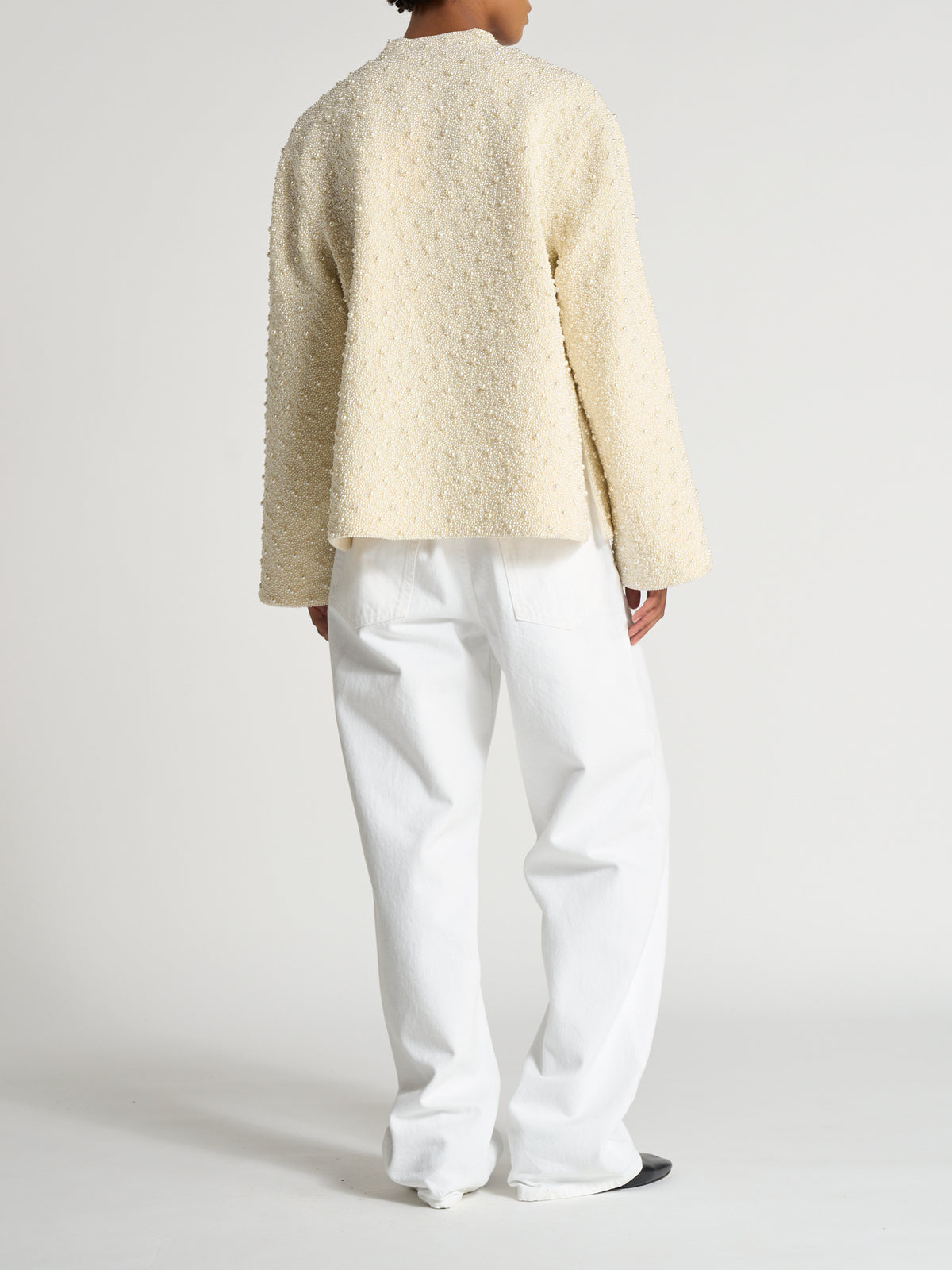 Back view of a model wearing the cream Hand-beaded pearl pankou jacket | pearl with white trousers in a minimalist studio setting
