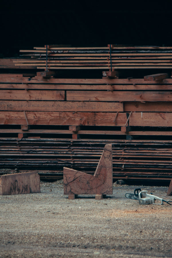 Brutalist Chair | 5788: A handcrafted oak chair featuring rough chainsaw cuts, showcasing its sculptural, functional design amidst a sawmill setting with wood planks and sawdust.