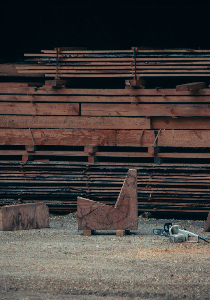 Brutalist Chair | 5788: A handcrafted oak chair featuring rough chainsaw cuts, showcasing its sculptural, functional design amidst a sawmill setting with wood planks and sawdust.