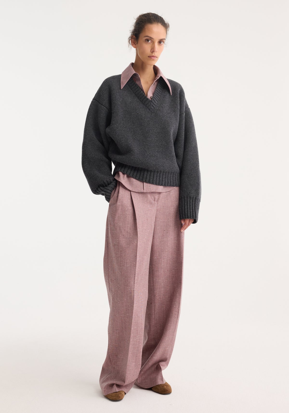 Model wearing a mid grey melange sculptural compact v-neck sweater layered over a pink collared shirt with loose pink trousers and brown shoes, standing against a plain background.