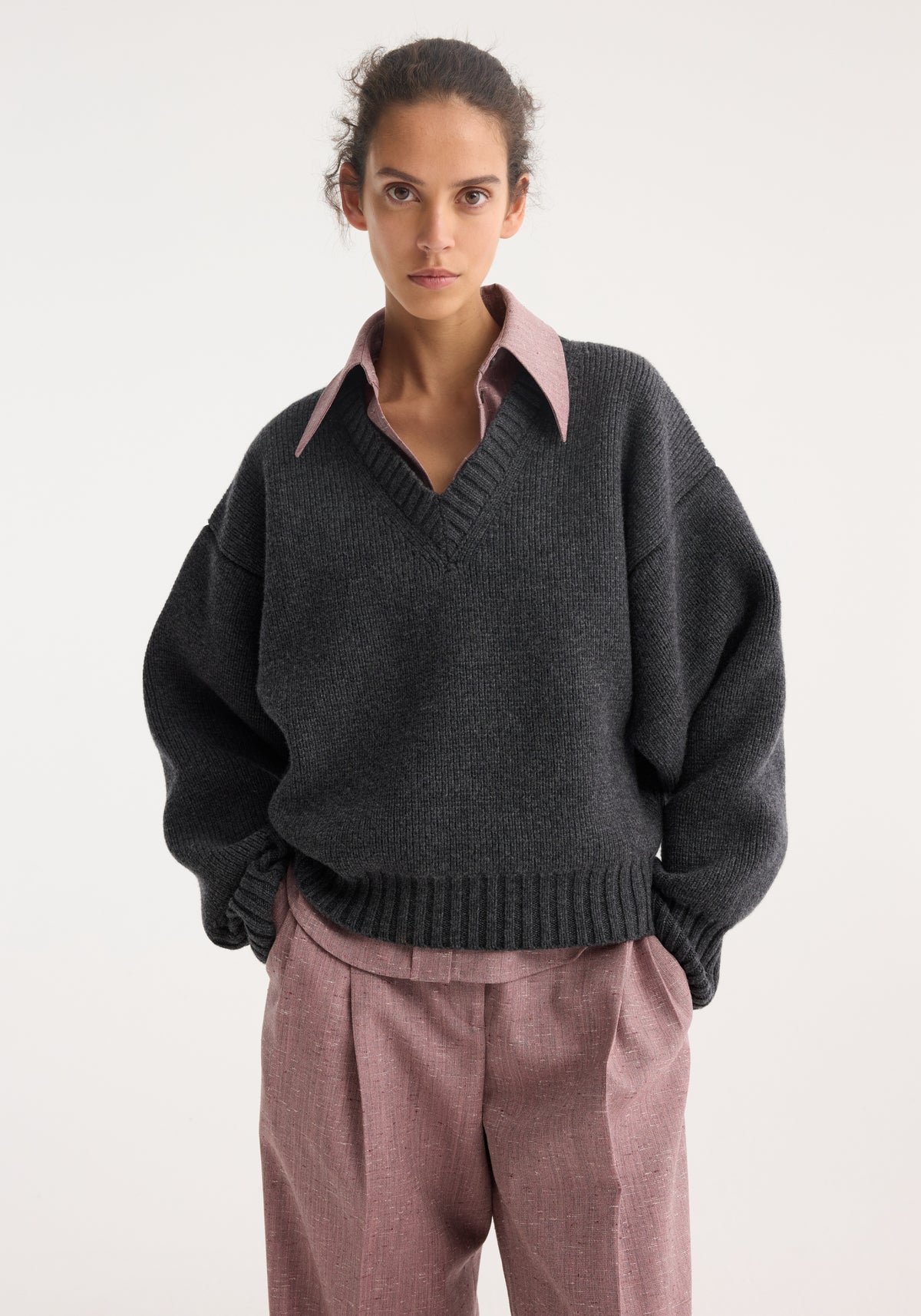 Model wearing a sculptural compact v-neck | mid grey melange knit sweater over a collared shirt with long sleeves and paired with loose pink trousers.