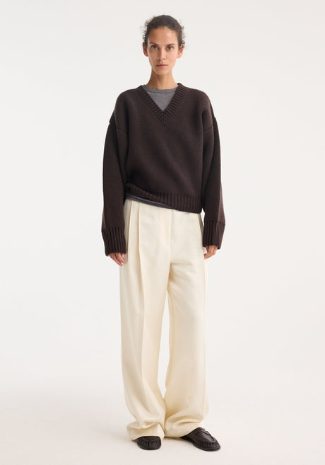 Model wearing Sculptural compact V-neck in dark brown, featuring a chunky knit design, relaxed fit, and Italian wool craftsmanship, paired with wide-leg cream trousers.