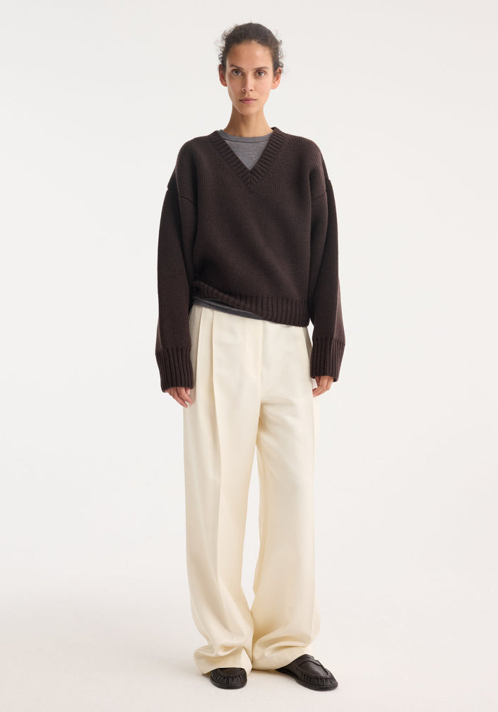 Model wearing Sculptural compact V-neck in dark brown, featuring a chunky knit design, relaxed fit, and Italian wool craftsmanship, paired with wide-leg cream trousers.