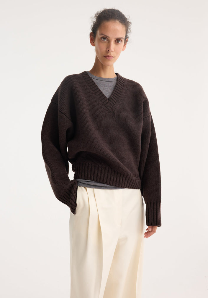 Model wearing Sculptural compact V-neck in dark brown, featuring a chunky knit design, relaxed fit, and Italian wool craftsmanship, paired with wide-leg cream trousers.