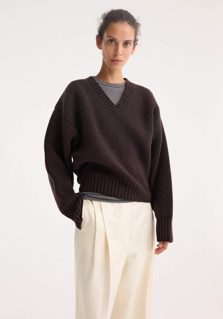 Model in oversized Sculptural compact v-neck dark brown sweater, layered over a grey top, paired with cream pleated trousers. Hand in pocket, facing forward.
