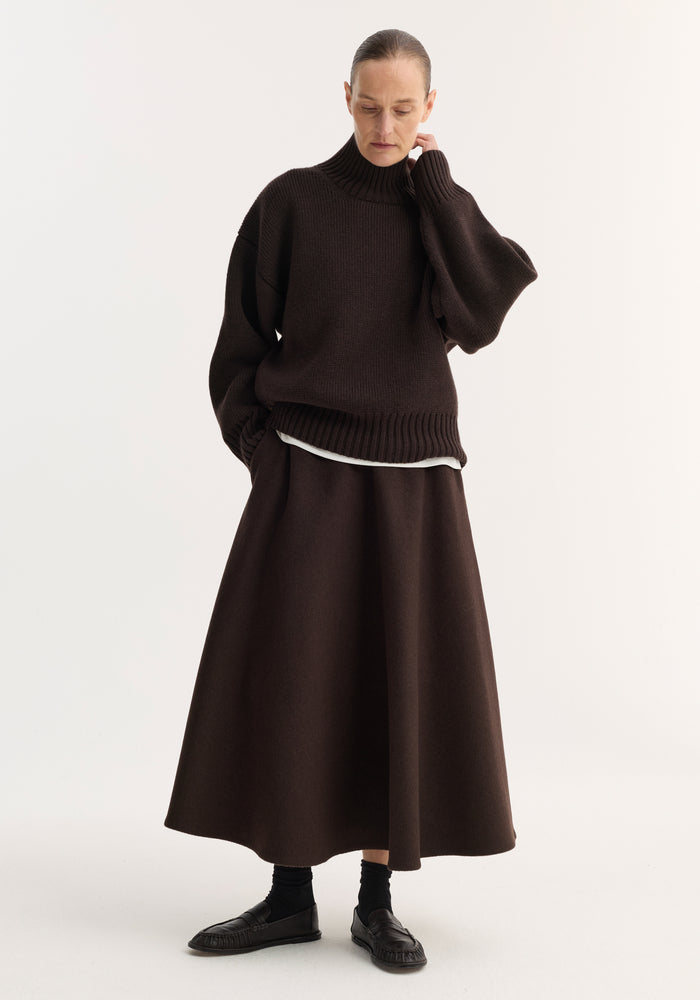 Person wearing a dark brown A-line skirt, crafted from a recycled wool blend, with side pockets and seamless zipper, paired with a knit turtleneck sweater.