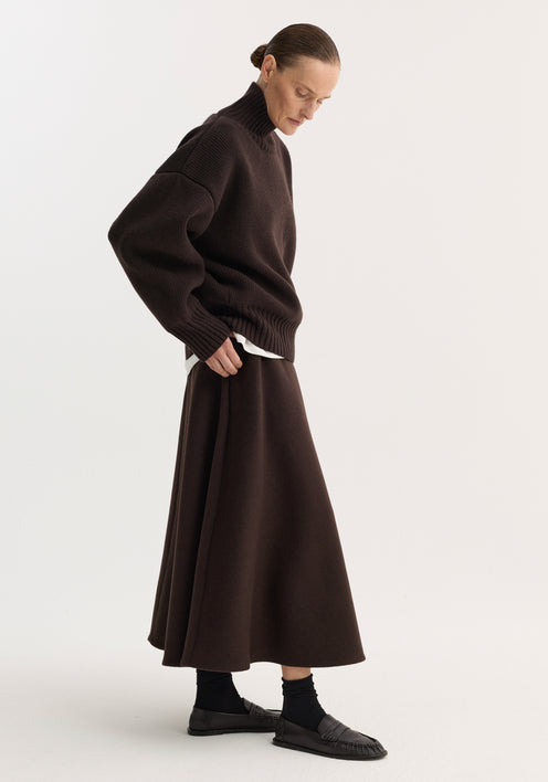 Person wearing a dark brown A-line skirt, crafted from a recycled wool blend, with side pockets and seamless zipper, paired with a knit turtleneck sweater.