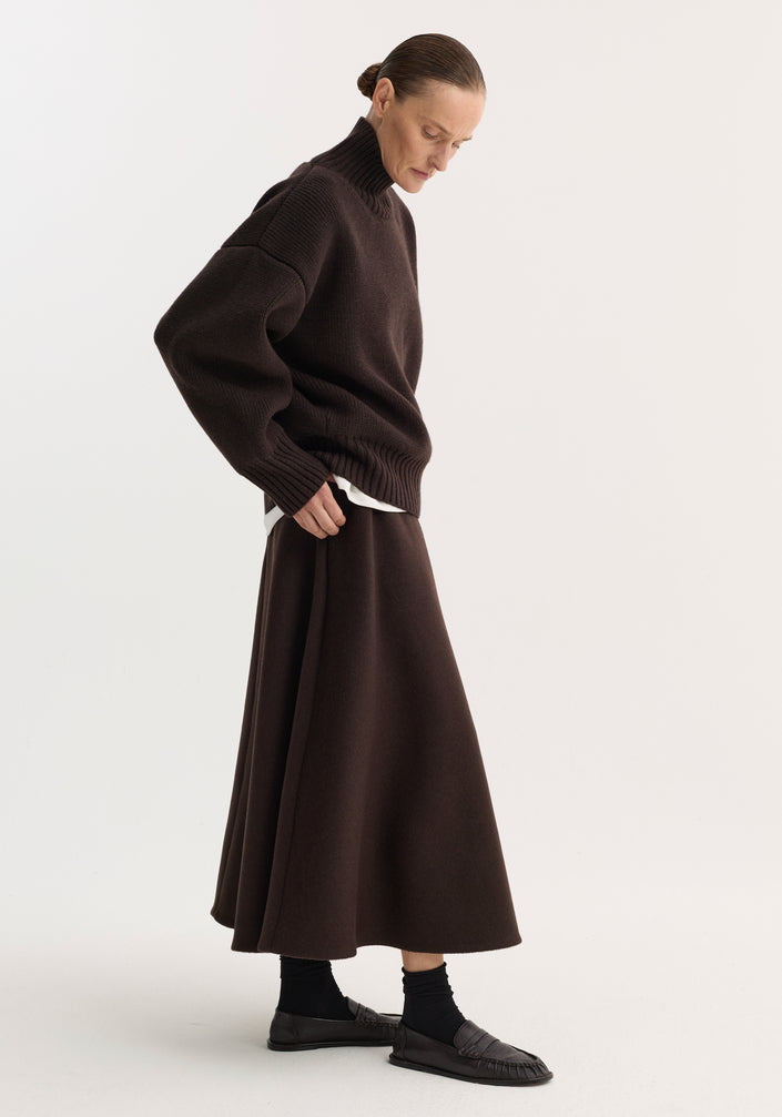 Person wearing a dark brown A-line skirt, crafted from a recycled wool blend, with side pockets and seamless zipper, paired with a knit turtleneck sweater.