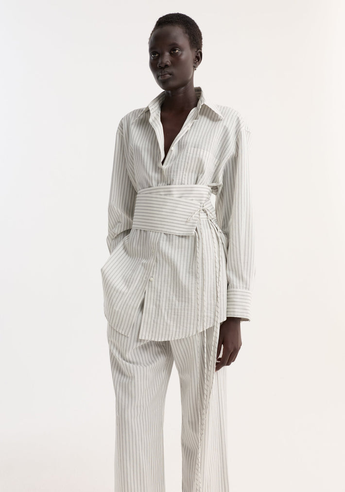 Model in Silk Stripe Belted Shirt with wide sleeves, matching belt, and pinstripe trousers, showcasing relaxed tailoring against a plain backdrop.