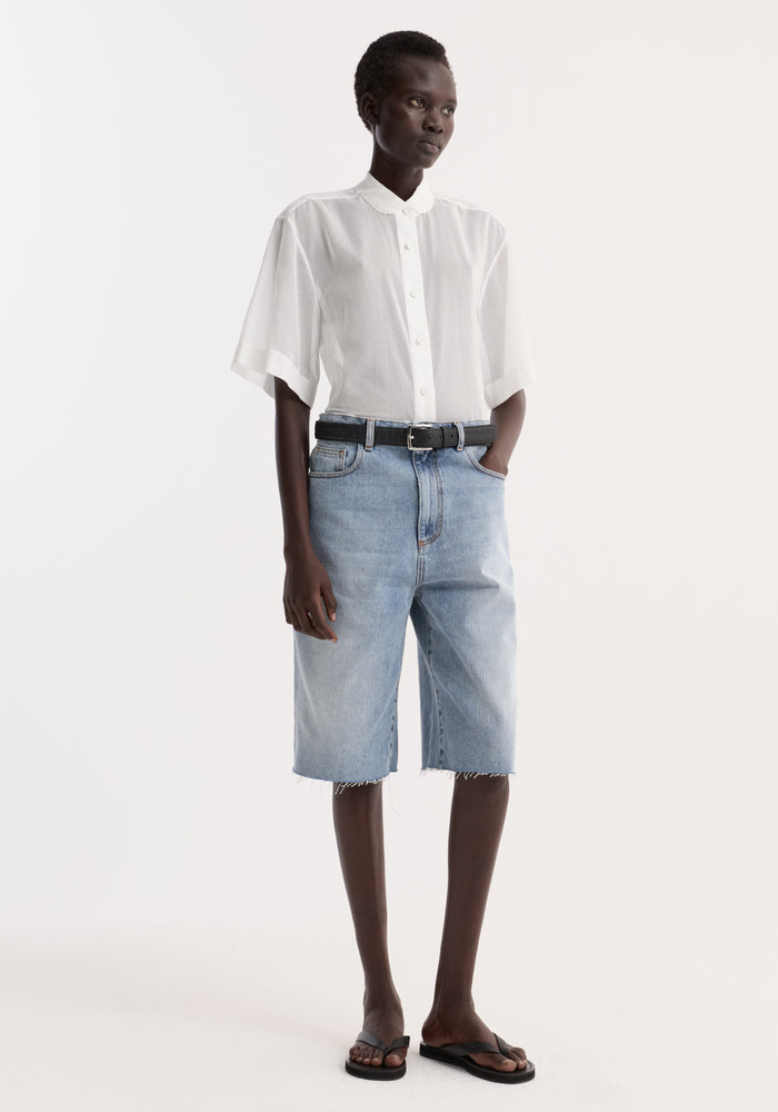 Model wearing an Embroidered muslin shirt in optic white with drop short sleeves, rounded collar, and light-wash denim bermuda shorts.
