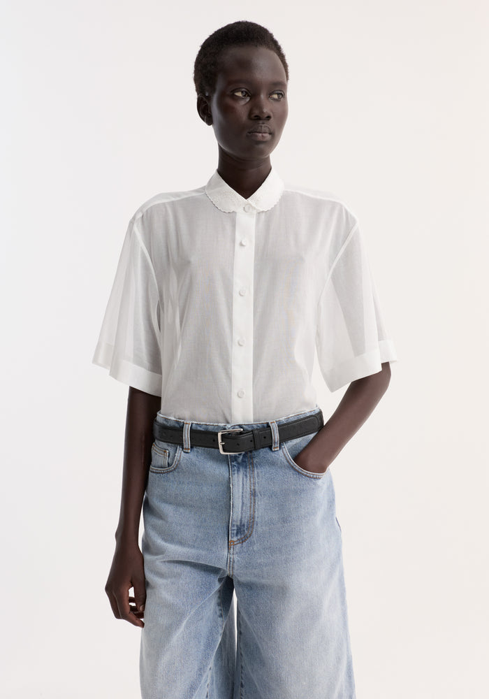 Person wearing an Embroidered muslin shirt | optic white, featuring a scalloped collar, short sleeves, and embroidery, tucked into light-wash denim with a black belt.