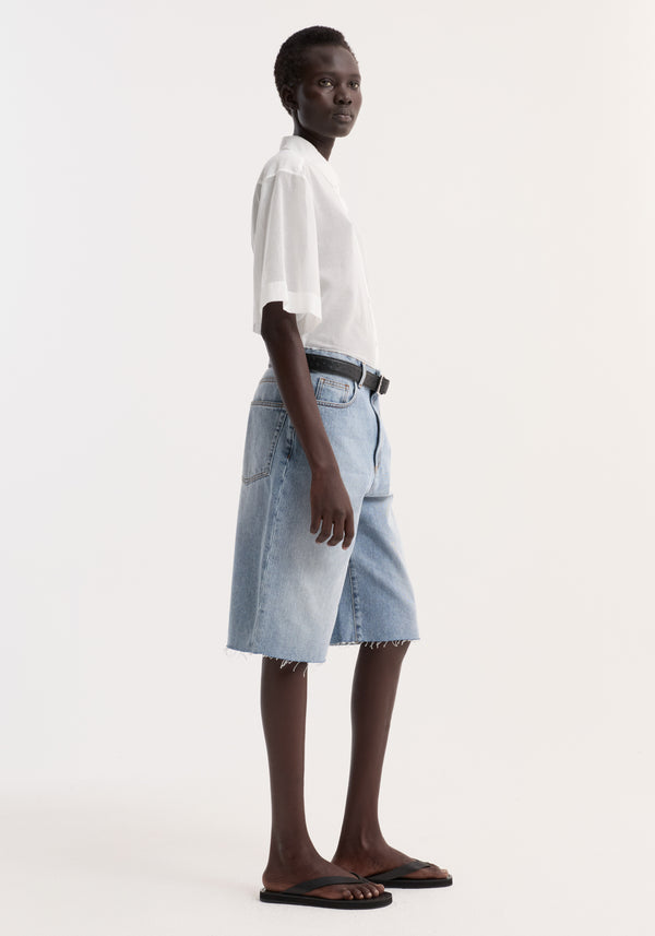 Model wearing an Embroidered muslin shirt in optic white, featuring sheer cotton, drop short sleeves, and subtle embroidery around the collar, paired with denim shorts.