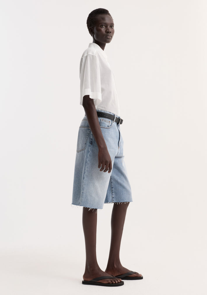 Model wearing an Embroidered muslin shirt in optic white, featuring sheer cotton, drop short sleeves, and subtle embroidery around the collar, paired with denim shorts.