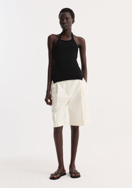 Model wears Chunky knit halter top | black, a sleeveless, hand-knit cotton design with ribbed hem and button closure, paired with pleated Bermuda shorts and flat sandals.