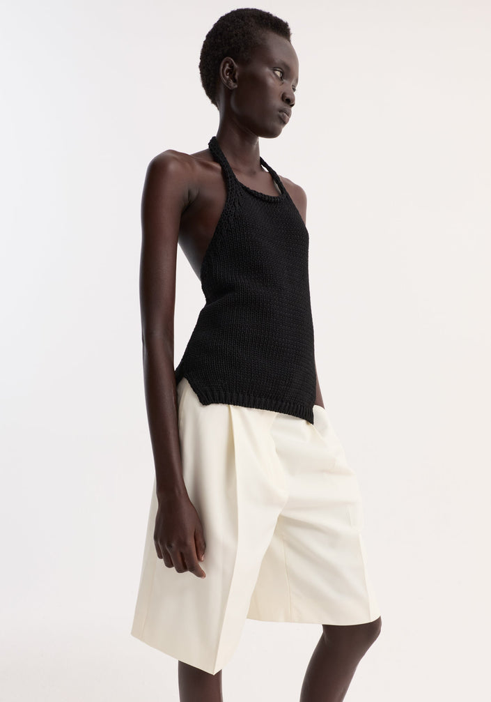 Model wears Chunky knit halter top | black, a sleeveless, hand-knit cotton design with ribbed hem and button closure, paired with pleated Bermuda shorts and flat sandals.