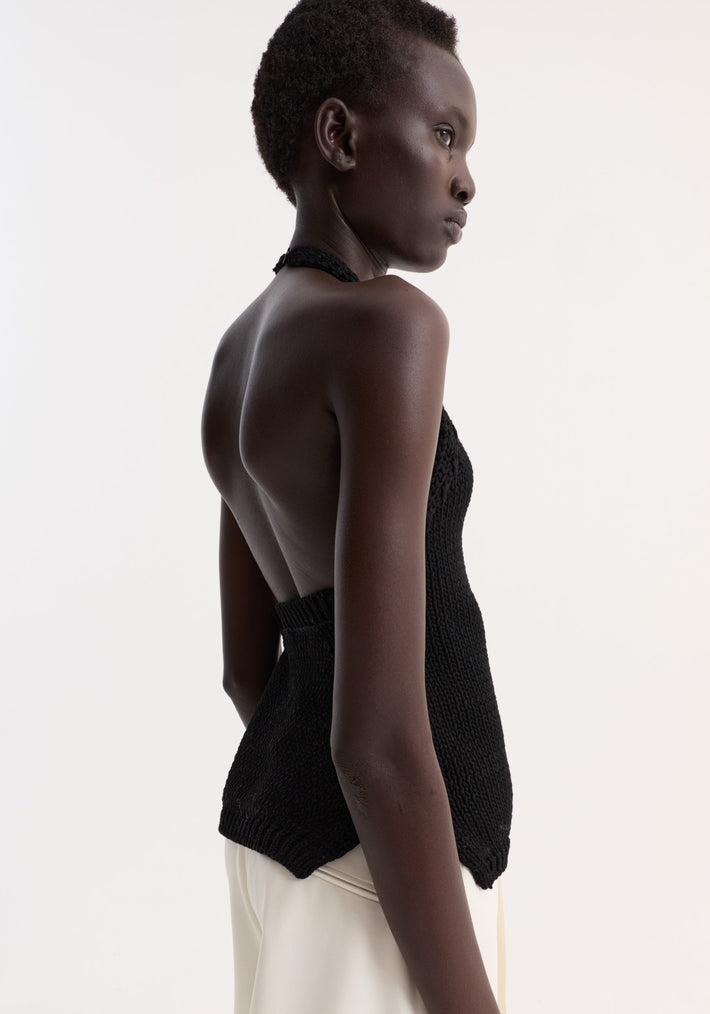 Side-profile of a model wearing a Chunky knit halter top | black, showcasing its open back, halter neckline, and button closure, paired with light trousers.