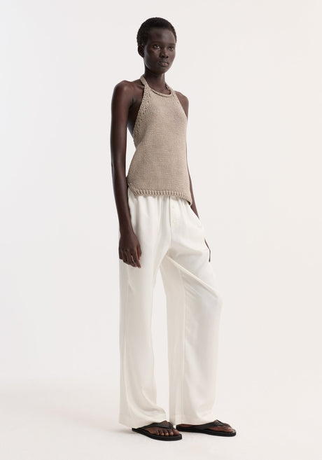 Model wearing Chunky knit halter top | stone, showcasing a sleeveless design with side slits, button neck closure, paired with white trousers and black sandals.