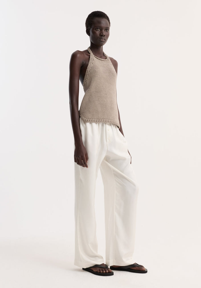 Model wearing Chunky knit halter top | stone, showcasing a sleeveless design with side slits, button neck closure, paired with white trousers and black sandals.