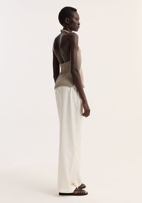 Model wearing Chunky knit halter top | stone, showcasing a sleeveless design with side slits, button neck closure, paired with white trousers and black sandals.