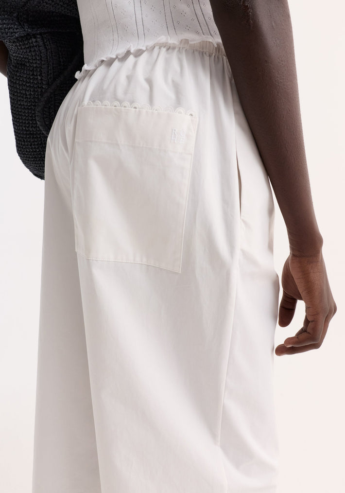Model wearing Embroidered poplin trousers in optic white, featuring a wide-leg silhouette, elasticated waistband, patch pocket with logo, and a black tote bag hint.
