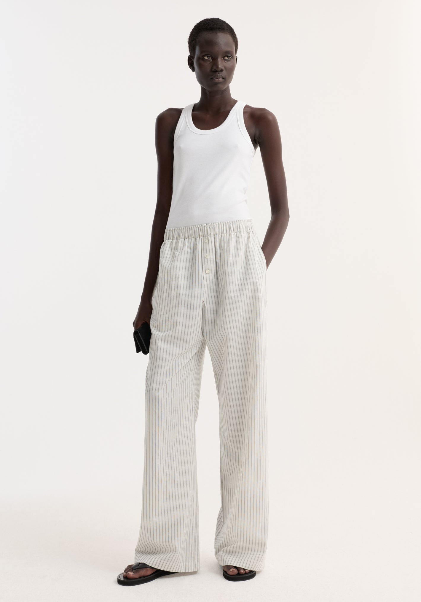 Silk stripe pyjama trousers with fine vertical stripes, wide-leg fit, and elasticated waistband, worn by a woman with black sandals and a clutch.