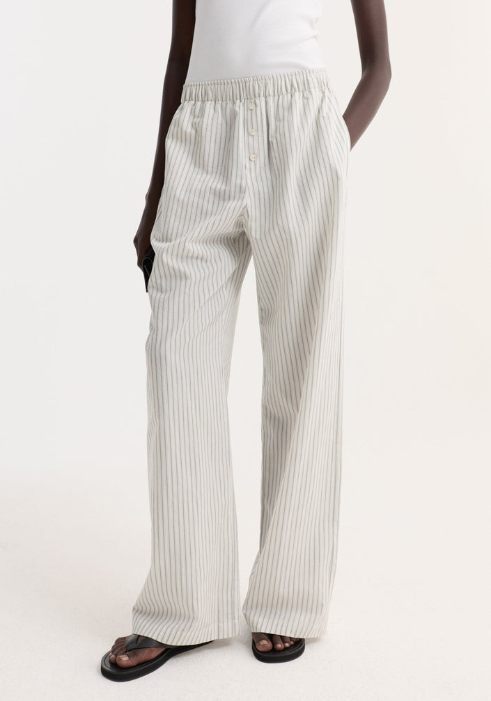 Person wearing Silk stripe pyjama trousers with fine vertical stripes, high-waisted, wide-leg design, and elasticated waistband, paired with a white tank top and black flip-flops.