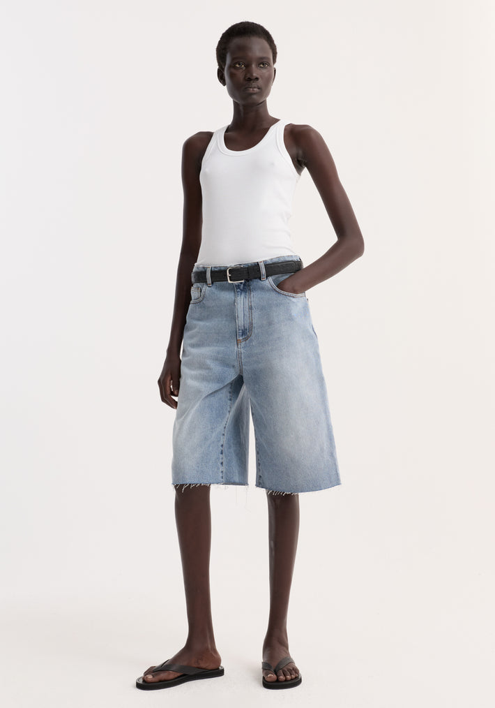 Baggy fit denim shorts in classic blue, featuring a raw hemline, modeled with a white rib-tank top and black flip-flops for a casual look.