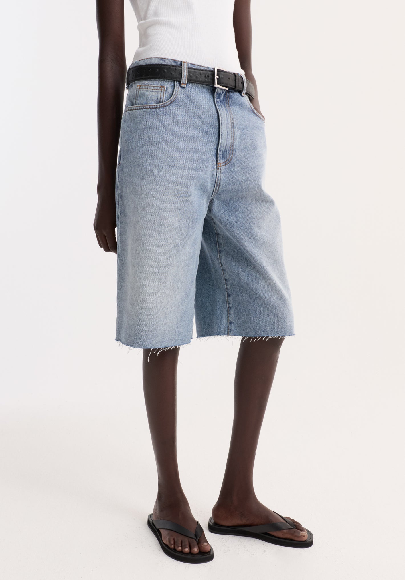 Baggy fit denim shorts in classic blue with a raw-edge hem, worn with a black belt and flip-flops, showcasing a relaxed, casual style.