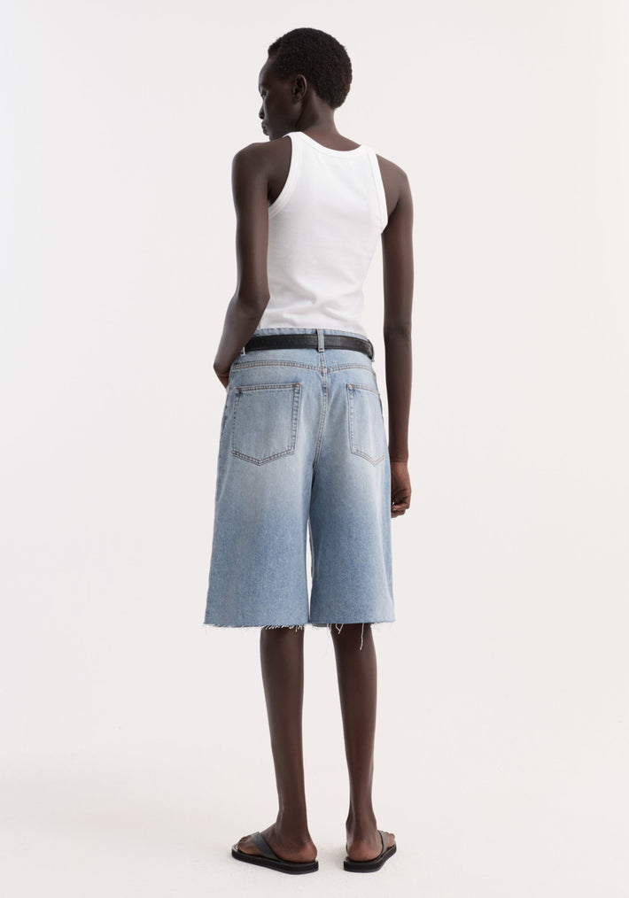 Baggy fit denim shorts in classic blue, featuring a raw hemline and knee-length cut, worn by a model with a white tank top and black slides.